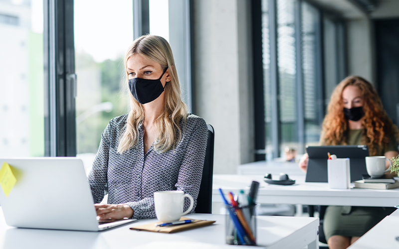 Woman sits at office desk with face mask on, with a colleague at a safe distance behind.