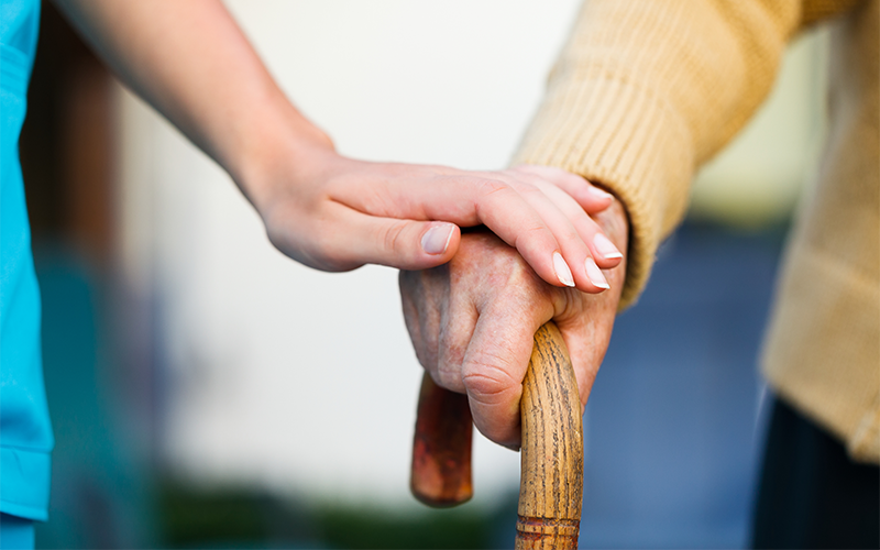Elderly man uses walking stick, assisted by a carer.